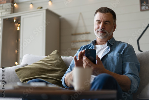 Happy smiling senior man using smartphone device while sitting on sofa at home