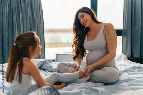 A pregnant mom puts her daughter's socks on while they sit on the bed and talk about their day