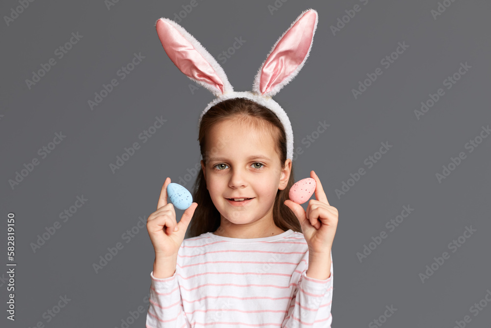 Portrait of adorable Caucasian little girl wearing bunny ears holding two Easter eggs, showing her artwork of painting testicles, looking at camera, isolated on gray background Portrait of adorable Caucasian little girl wearing bunny ears holding two Easter eggs, showing her artwork of painting testicles, looking at camera, isolated on gray background