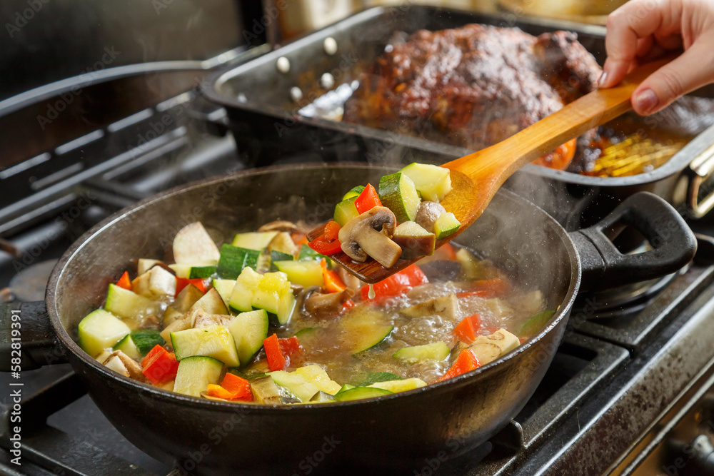 A frying pan with stewed vegetables and a wooden spoon on a stove next to a tray with baked chicken. A frying pan with stewed vegetables and a wooden spoon on a stove next to a tray with baked chicken.