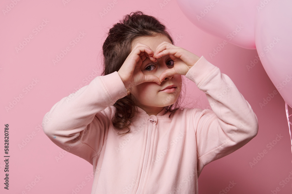 Beautiful Caucasian little child girl looking through a heart made from her fingers, gesturing with hand sign, showing you her affection, sympathy and love, isolated over pink colored background Beautiful Caucasian little child girl looking through a heart made from her fingers, gesturing with hand sign, showing you her affection, sympathy and love, isolated over pink colored background