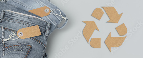 Stack of gray jeans with empty tags. Long panoramic banner with circular fashion, eco friendly sustainable shopping, donation, thrift stores concept. Top view over womens clothes and recycling symbol photo