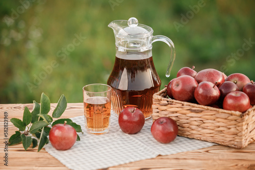 A glass with apple juice and basket with apples on wooden table with natural orchard background. Vegetarian fruit composition