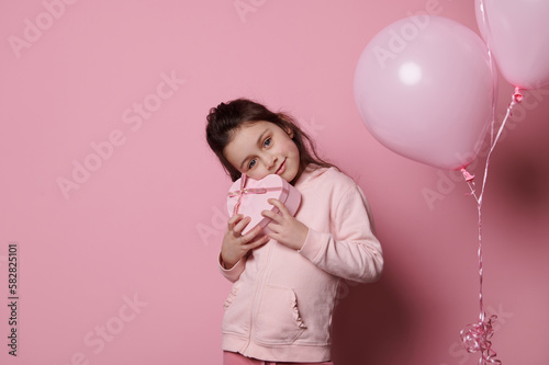 Beautiful Caucasian little girl, enjoying festive occasion event, holding a cute heart shaped gift box, smiling, isolated on pink color background with pink pastel air balloons. Mother's Day concept photo