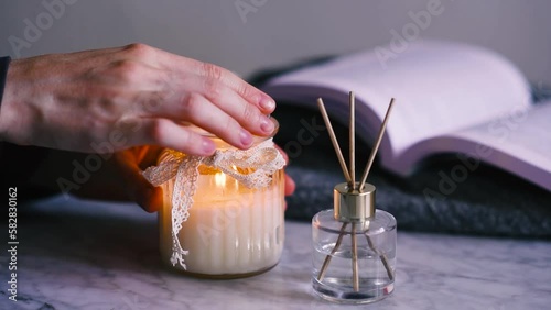 Woman hands with burning scented candle with home liquid diffuser over open book in room closeup. Cozy atmosphere.