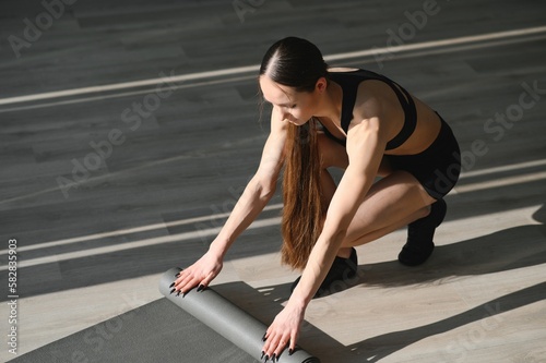 Time for meditation, fitness session, well-being concept. Girl wearing grey sporty pants rolling fitness mat before, after class in yoga studio club or at home on wooden floor.