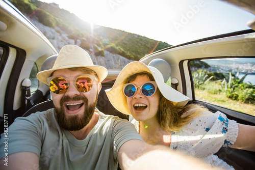 Romantic couple making selfie on smartphone camera in rental cabrio car on ocean or sea beach enjoying summer vacation together and taking picture on cellular resting near sea on weekends