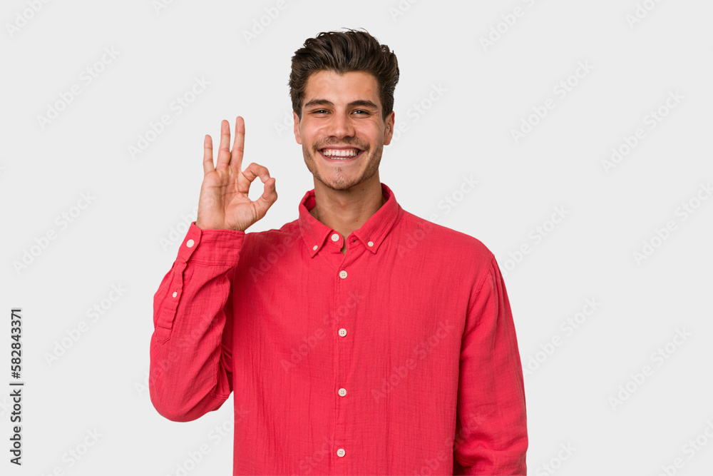 Young handsome caucasian man isolated on white background winks an eye and holds an okay gesture with hand. Young handsome caucasian man isolated on white background winks an eye and holds an okay gesture with hand.