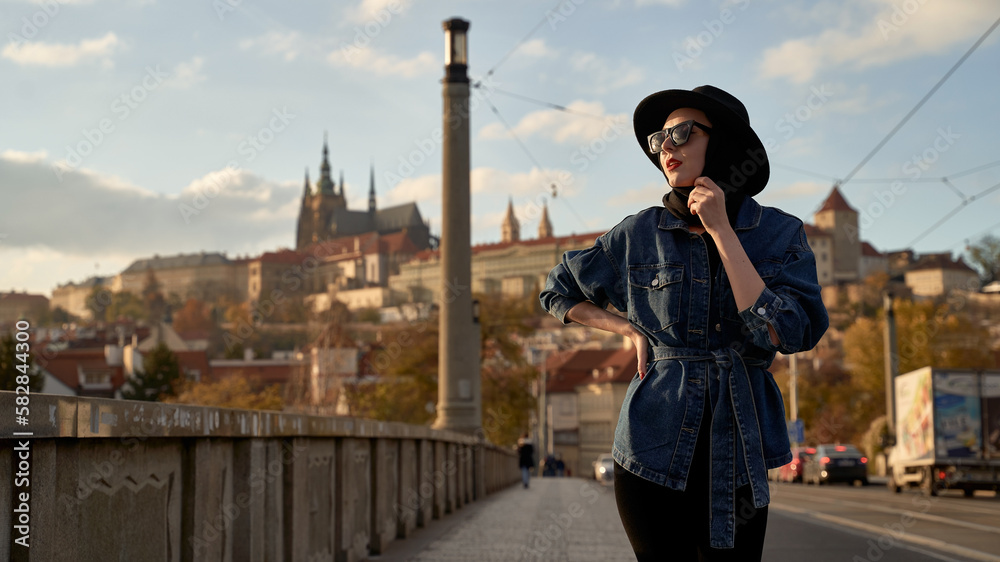 Stylish beautiful young woman earing black hat in Prague on a bridge with Prague Castle or Prazsky Hrad on background. Elegant retro lady fine art portrait.