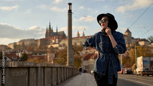 Obraz na płótnie Stylish beautiful young woman earing black hat in Prague on a bridge with Prague Castle or Prazsky Hrad on background. Elegant retro lady fine art portrait. w salonie