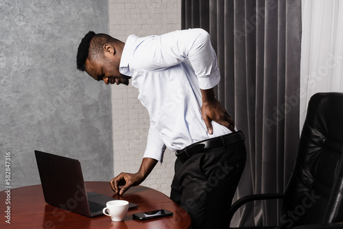 Cervical spine osteochondrosis is radicular syndromes of african american man. African manholds his back and feels pain in his back and neck after working for long hours with laptop online in office. photo