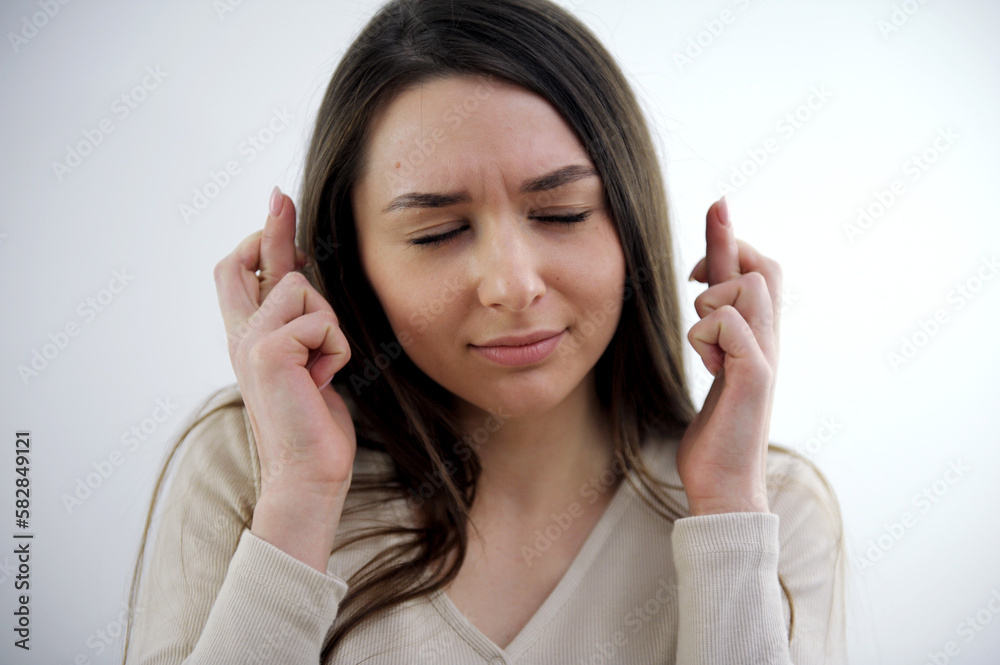Pleased attractive European woman makes wish to win, raises hands with crossed fingers, waits for lottery results, closes eyes, dressed in fashionable dress, isolated over white wall