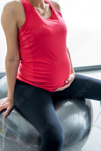 Pregnant woman in a vibrant sports shirt sitting on an exercise ball and holding her hands on her belly. Pregnancy, maternity, motherhood, sport, and health concept. Close-up with a bright background.
