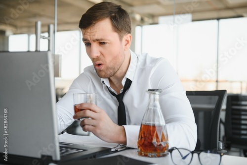Young stressed handsome businessman working at desk in modern office shouting at laptop screen and being angry about financial situation, jealous of rival capabilities, unable to meet client needs