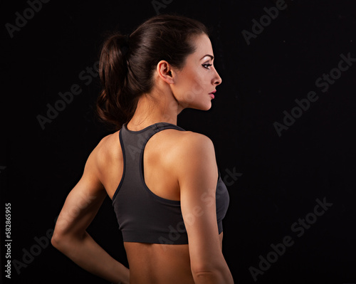Serious female sporty muscular with ponytail doing stretching workout the shoulders, blades and arms in sport bra on black background with empty space. Back view, profile face, healthy posture.