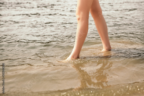 Close up naked slim woman legs walking barefoot along the river. Female feet on sea beach with splashing waves and foam. Girl tourist on vacation. Lady go on shore after bathing
