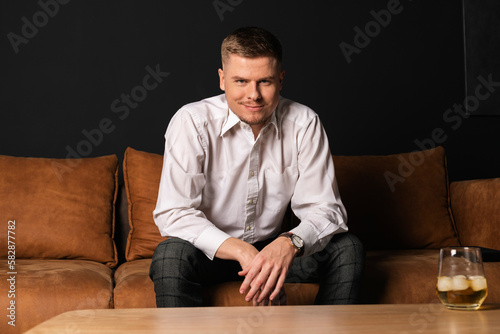 An attractive young successful man in a white shirt sits on a brown couch and drinks whiskey with ice. A happy smiling businessman.