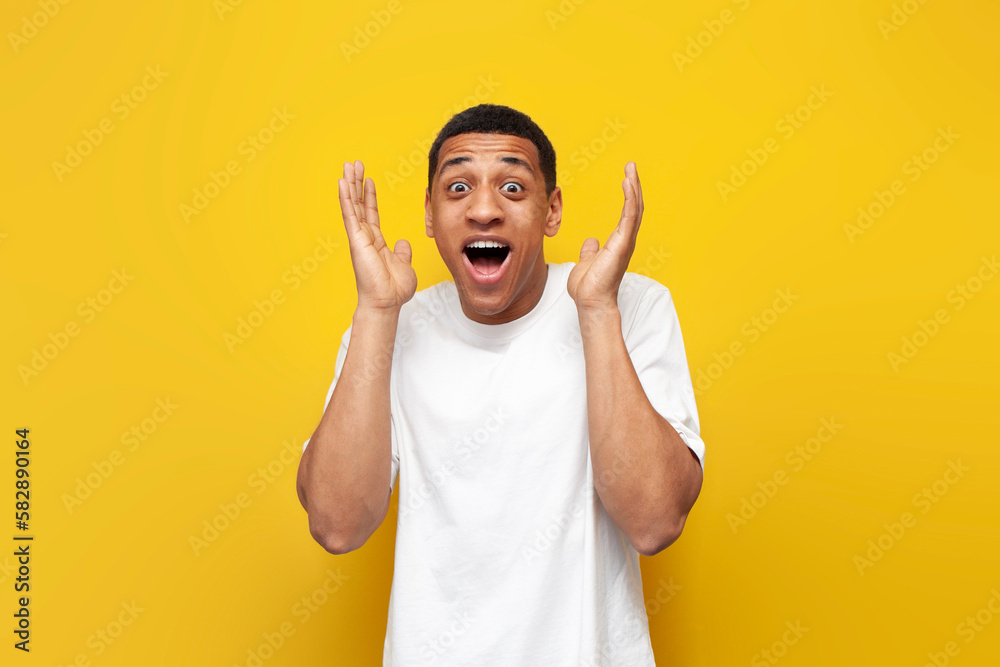 young surprised guy african american in white t-shirt raises his hands in front of him and opens his mouth - obrazy, fototapety, plakaty 