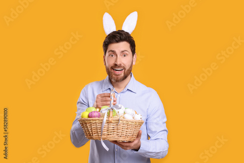 Portrait of happy man in cute bunny ears headband holding wicker basket with Easter eggs on orange background