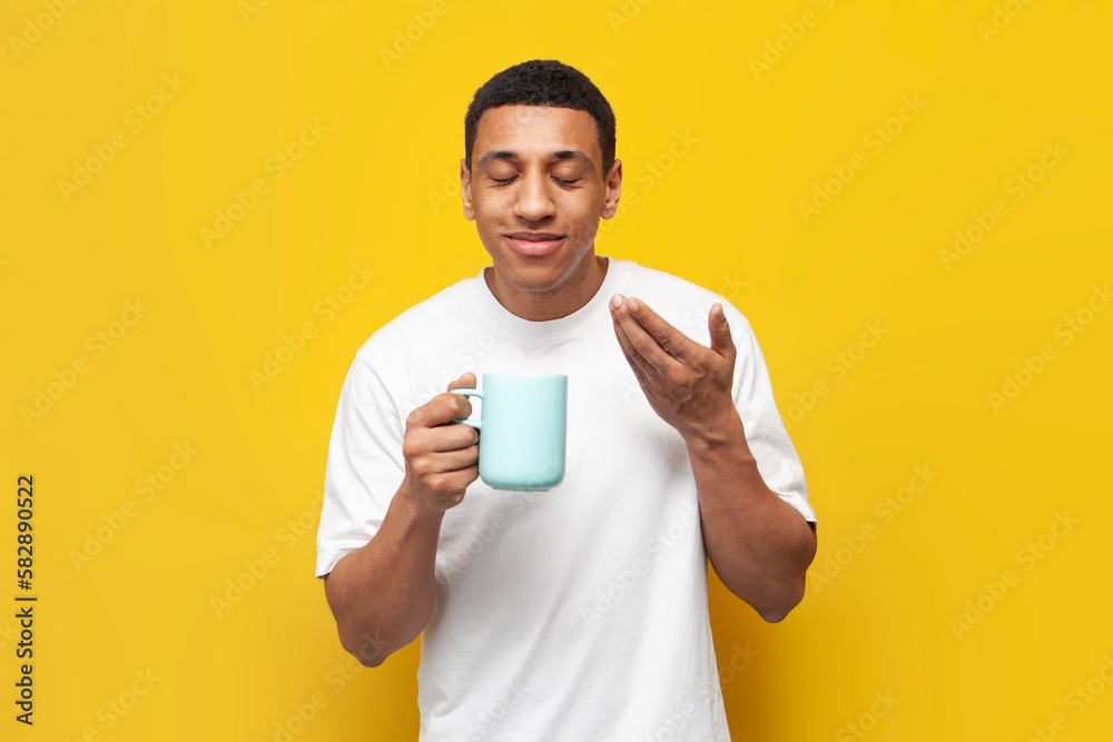 young african american guy in white t-shirt holds cup of coffee and smells the aroma on yellow isolated background young african american guy in white t-shirt holds cup of coffee and smells the aroma on yellow isolated background