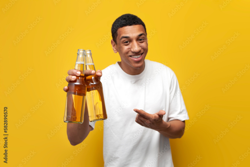 young african american guy in white t-shirt holds two bottles of beer and offers drink on yellow isolated background young african american guy in white t-shirt holds two bottles of beer and offers drink on yellow isolated background
