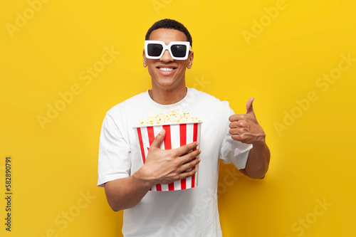 young african american guy in 3d glasses watches movie with popcorn and showing like on yellow isolated background photo