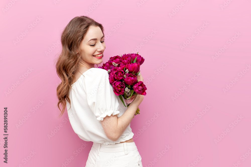 young attractive woman in white festive clothes holding bouquet of pink tulips on pink isolated background
