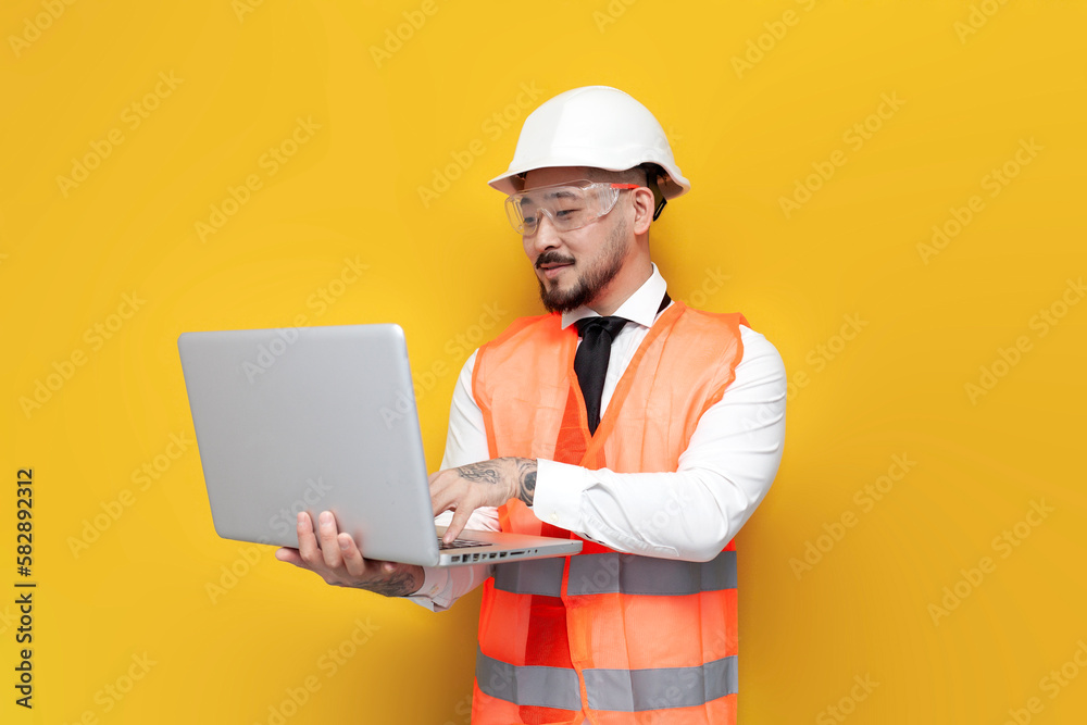 asian foreman in uniform uses laptop on yellow isolated background, korean civil engineer in hard hat and glasses asian foreman in uniform uses laptop on yellow isolated background, korean civil engineer in hard hat and glasses