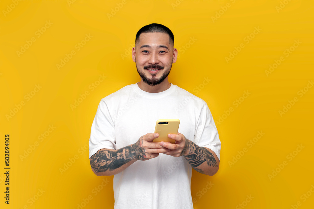 adult tattooed asian man in white t-shirt uses smartphone on yellow isolated background and smiles adult tattooed asian man in white t-shirt uses smartphone on yellow isolated background and smiles