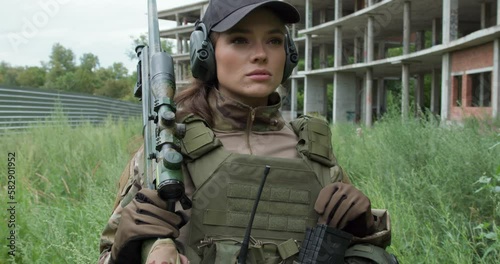 Young serious confident woman in military uniform armed with rifle looking sideway, girl with gun on her shoulder pretecting area, territory ca,mera motion security ruined biuilding in the background photo