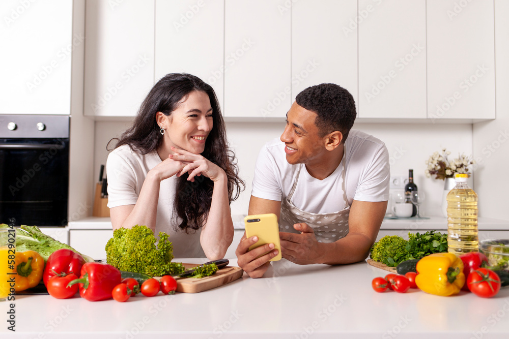 young multiracial couple preparing veggie salad of vegetables and herbs and using smartphone