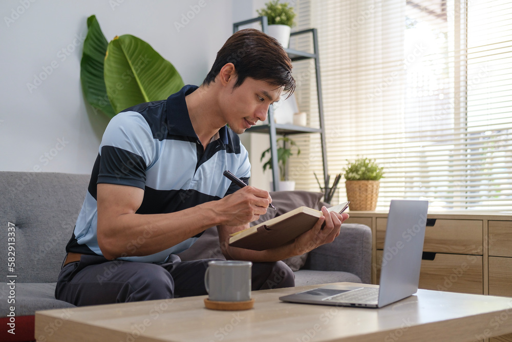 Shot of adult asian man using a laptop and making notes on notebook while relaxing on a couch in at home.