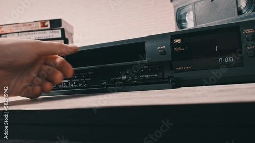 Insert VHS cassette into VCR player. Black vintage videotape cassette recorder on a desk with many archived video cassettes. Male hand inserting old VHS Tape into a retro player. Home video, nostalgia photo