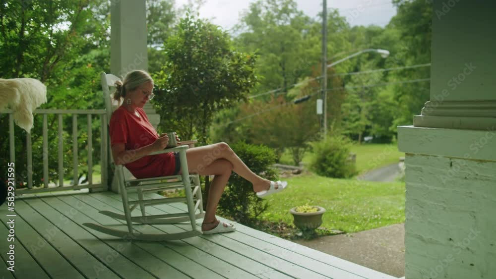 Young woman sitting on a rocking chair on her front porch, relaxing and drinking tea (wide)