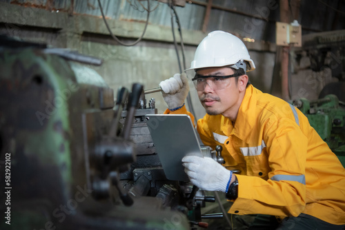 Asian engineer wearing yellow work clothes white hardhat and wear glove looking and touch to the machine while holding tablet glasses, Industry and production concept.