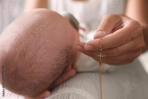 Mother holding Christian cross near newborn baby indoors, closeup