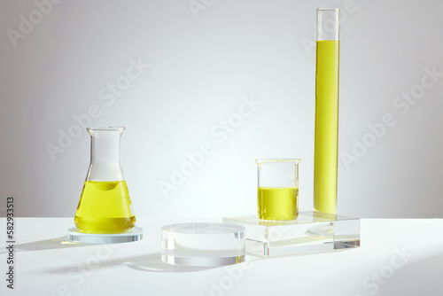 Laboratory concept with erlenmeyer flask, beaker and test tube containing yellow liquid and empty transparent podium on light background. Background for the presentation of cosmetic products
