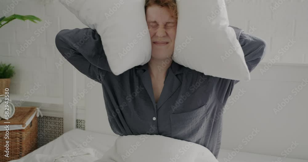 Portrait of annoyed young woman in pajamas covering ears with pillows