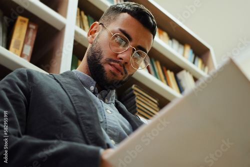 Young bearded man with eyeglasses reading book while standing by bookcase in library, flipping pages, enjoying literature. Low angle reader going through interesting plot. Concept of education
