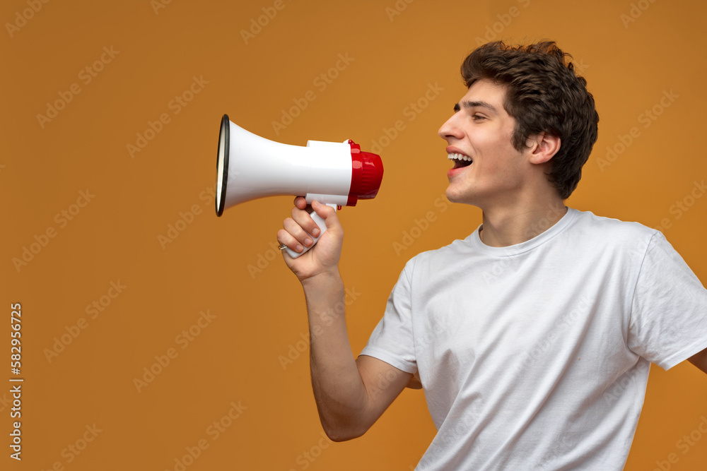 Young man shouting into megaphone making announcement against yellow background Young man shouting into megaphone making announcement against yellow background