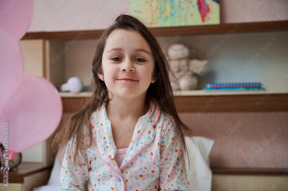 Caucasian lovely little child girl in elegant white pajamas with colorful dots, smiling broadly looking at camera, sitting on the bed in her glamour bedroom, decorated with pink helium balloons Caucasian lovely little child girl in elegant white pajamas with colorful dots, smiling broadly looking at camera, sitting on the bed in her glamour bedroom, decorated with pink helium balloons