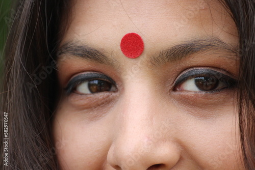 Left Eye in Focus, burred effect, an Indian woman closeup looking into camera. She is wearing red color Bindi on her forehead and having a soft smile on the face.  photo