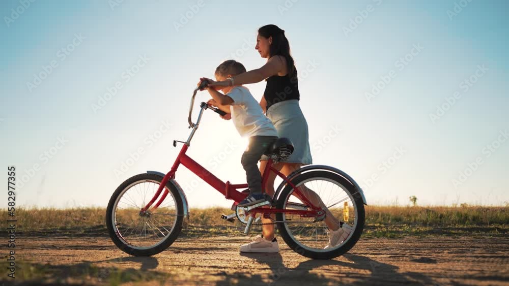 Happy family concept. Mom teaches her son to ride bike on green grass. Child rides bicycle along rural road. Green energy. Mom teaches her son to ride bike for the first time in park at sunset. Happy family concept. Mom teaches her son to ride bike on green grass. Child rides bicycle along rural road. Green energy. Mom teaches her son to ride bike for the first time in park at sunset.