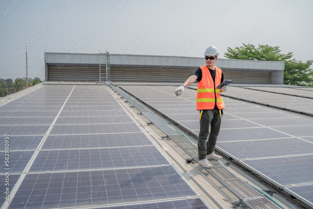 Asian engineers wearing protective vest and white hardhat standing holding tablet pointing to solar cell roof with copy space, Photovoltaic technology concept.