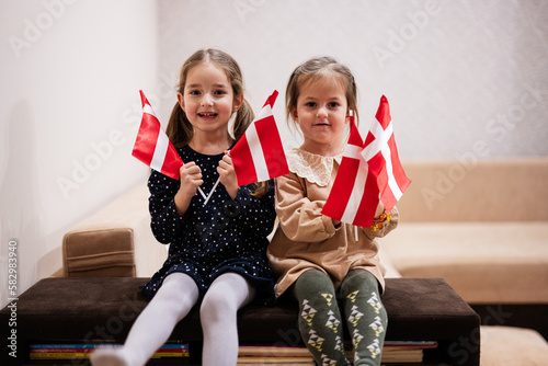 Two sisters are sitting on a couch at home with danish flags on hands. Denmark children girls with flag .