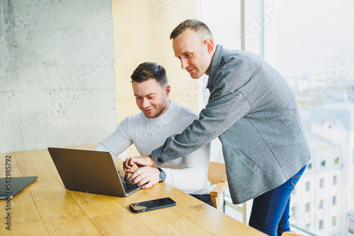 Two male colleagues while working with a laptop in the office. Two goal-oriented entrepreneurs collaborate in a modern workspace. Two young businessmen work in a bright office