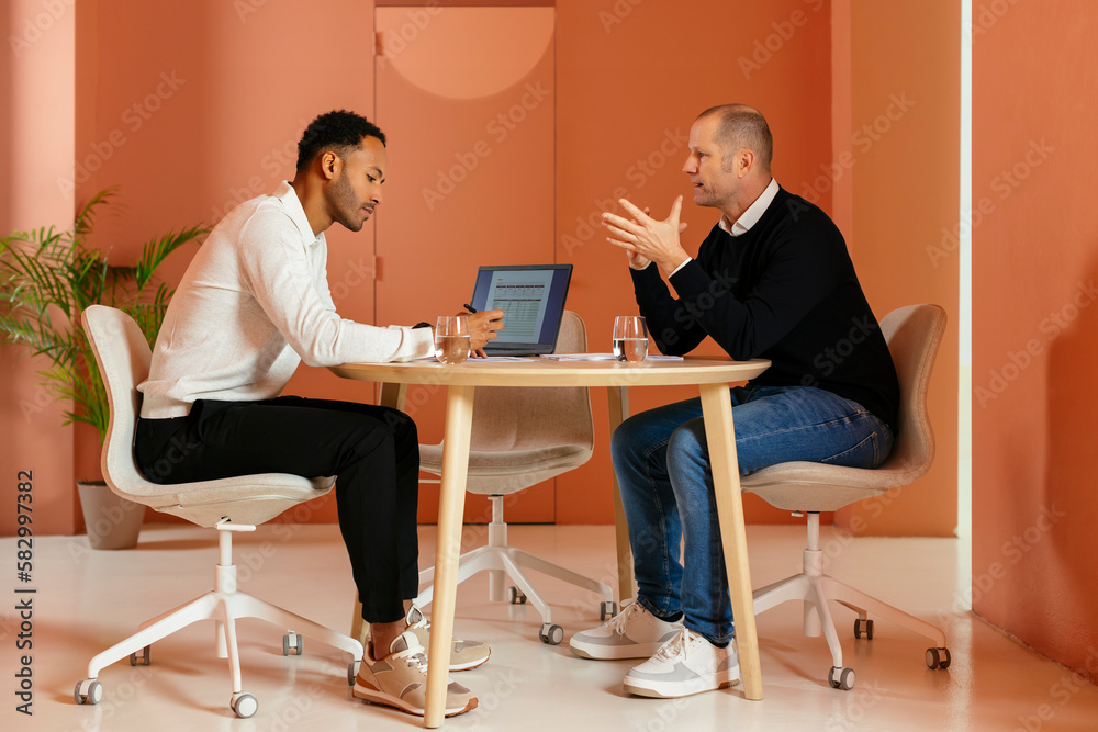 Businessman discussing with colleague at desk in front of orange wall Businessman discussing with colleague at desk in front of orange wall