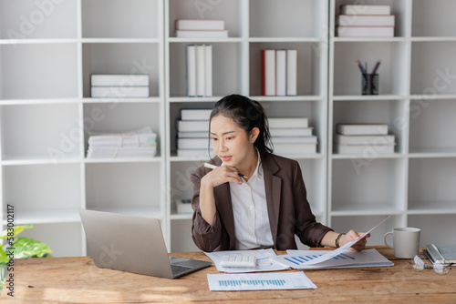 Young beautiful asian businesswoman using laptop and smart phone and calculator for doing financial mathematics on desk, tax, report, accounting, statistics and analytical research concept.