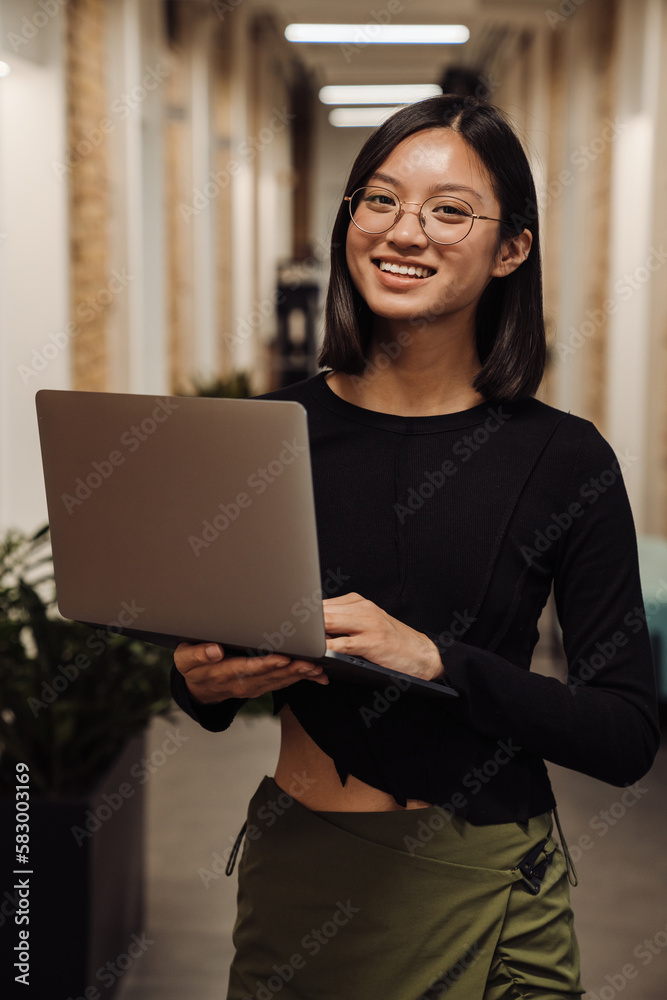 Smiling asian business woman working on laptop while standing in office Smiling asian business woman working on laptop while standing in office