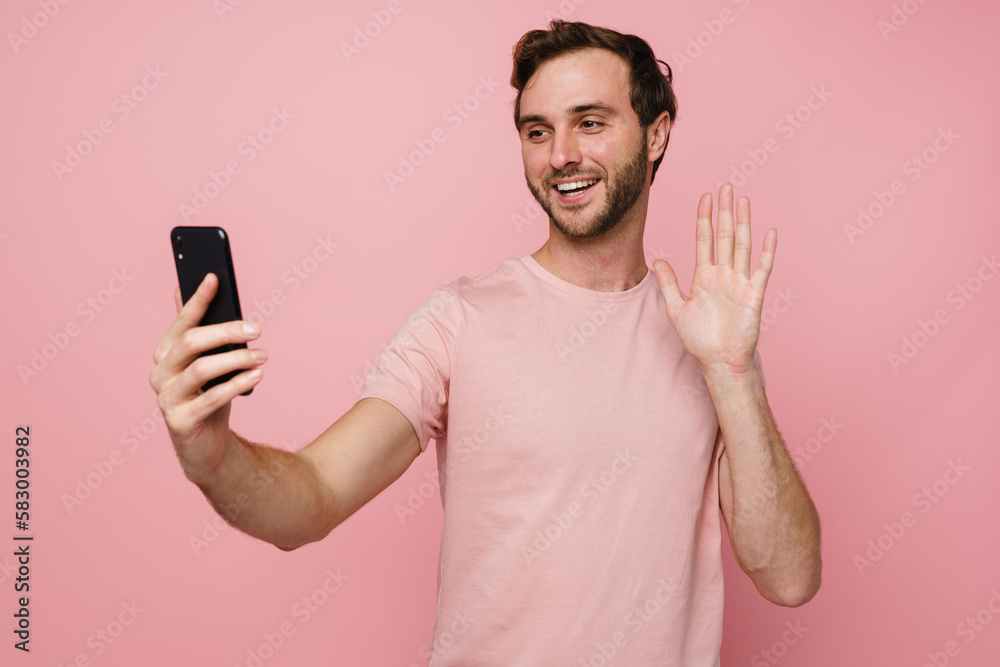 Positive man waving hand while taking selfie on mobile phone isolated over pink wall Positive man waving hand while taking selfie on mobile phone isolated over pink wall